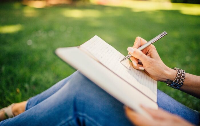Nainen istuu nurmikolla ja kirjoittaa sylissään olevaan muistikirjaan.