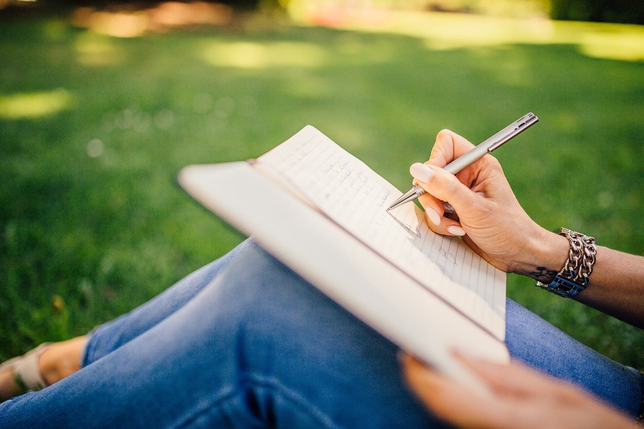 Nainen istuu nurmikolla ja kirjoittaa sylissään olevaan muistikirjaan.
